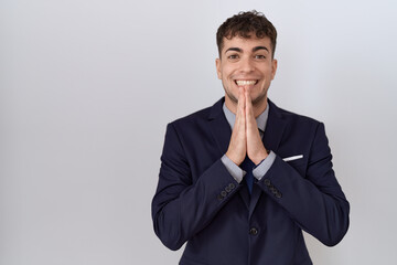 Young hispanic business man wearing suit and tie praying with hands together asking for forgiveness smiling confident.