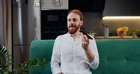 Attaractive man sit in kitchen hold smart phone talk on speakerphone. Male recording audio message...
