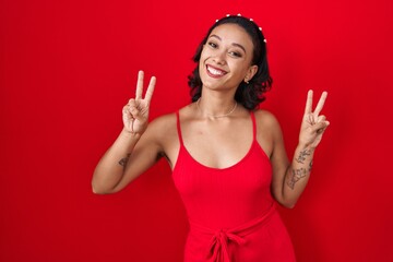 Young hispanic woman standing over red background smiling looking to the camera showing fingers doing victory sign. number two.