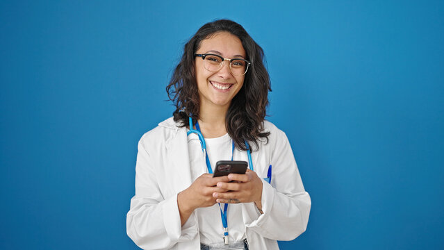 Young beautiful hispanic woman doctor smiling using smartphone over isolated blue wall background