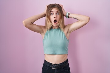 Obraz na płótnie Canvas Blonde caucasian woman standing over pink background crazy and scared with hands on head, afraid and surprised of shock with open mouth