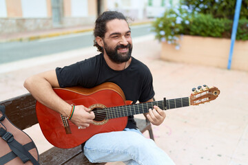 Young hispanic man musician playing classical guitar sitting on bench at park