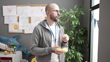 Young bald man working as teacher eating chips potatoes at kindergarten