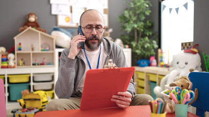 Young bald man preschool teacher reading document speaking on the phone at kindergarten
