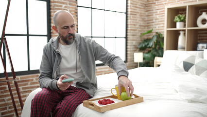 Young bald man having breakfast using smartphone at bedroom