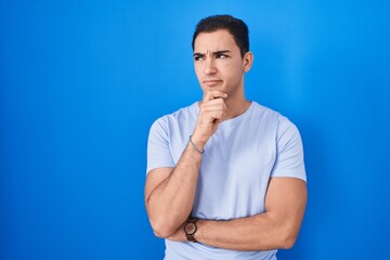 Fototapeta premium Young hispanic man standing over blue background thinking worried about a question, concerned and nervous with hand on chin