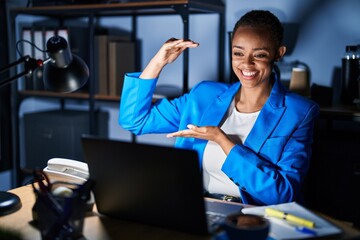 Beautiful african american woman working at the office at night gesturing with hands showing big and large size sign, measure symbol. smiling looking at the camera. measuring concept.
