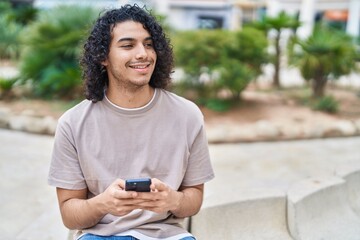 Young latin man using smartphone sitting on bench at park