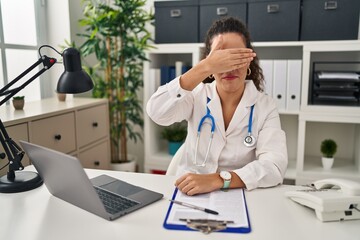 Young hispanic woman wearing doctor uniform and stethoscope covering eyes with hand, looking...