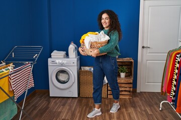 Young beautiful hispanic woman smiling confident holding basket with clothes at laundry room