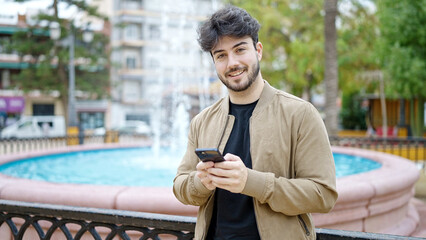 Young hispanic man using smartphone smiling at park