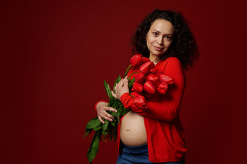 Multi ethnic beautiful pregnant woman, wearing red shirt, holding a cute bouquet of tulips, posing bare belly on isolated color background. Expectant mother. Maternity. Happy Women's and Mother's Day