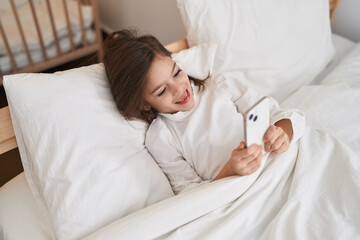 Adorable hispanic girl using smartphone lying on bed at bedroom