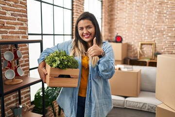 Young hispanic woman moving to a new home holding plants smiling happy and positive, thumb up doing excellent and approval sign