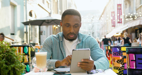 Front view of handsome busy high-skilled young black-skinned man in casual clothes which working on tablet pc while sitting in cozy street cafe at time break
