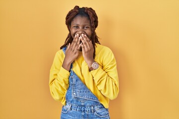 African woman standing over yellow background laughing and embarrassed giggle covering mouth with...