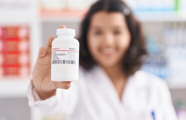 Young woman pharmacist smiling confident holding pills bottle at pharmacy