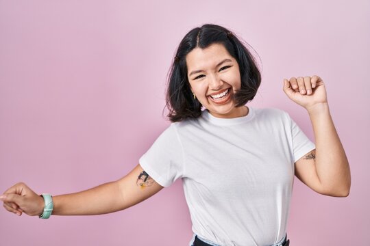 Young hispanic woman wearing casual white t shirt over pink background dancing happy and cheerful, smiling moving casual and confident listening to music