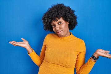Black woman with curly hair standing over blue background clueless and confused expression with arms and hands raised. doubt concept.