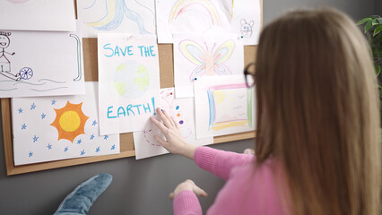 Young blonde woman working as teacher hanging save the earth draw on corkboard at kindergarten