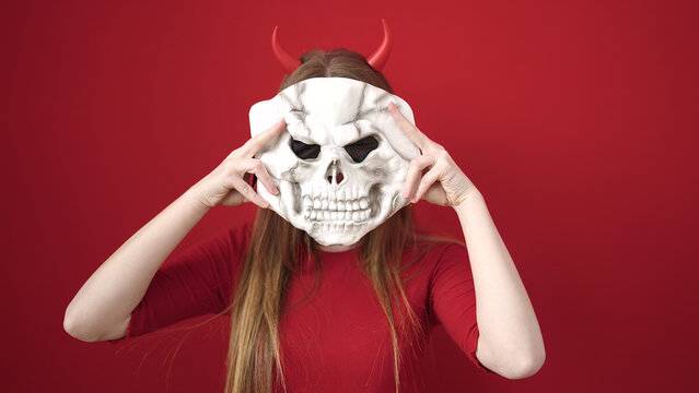 Young blonde woman wearing devil costume holding skull mask over isolated red background - Powered by Adobe