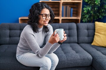 Young latin woman drinking coffee sitting on sofa at home