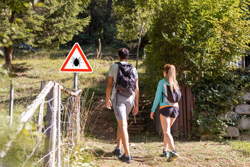 Man and woman hiking in Infected ticks forest with warning sign. Risk of tick-borne and lyme...