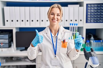 Young caucasian woman working at scientist laboratory holding test tubes smiling happy and positive, thumb up doing excellent and approval sign