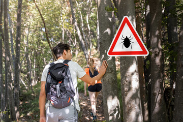 Man and woman hiking in Infected ticks forest with warning sign. Risk of tick-borne and lyme...