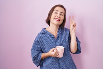 Middle age hispanic woman drinking a cup coffee smiling looking to the camera showing fingers doing victory sign. number two.