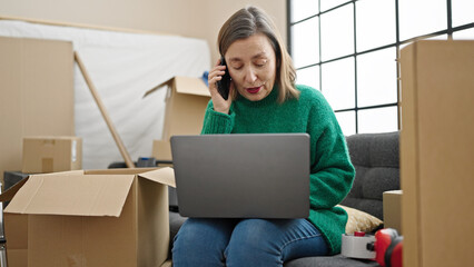 Mature hispanic woman with grey hair talking on smartphone using laptop at new home