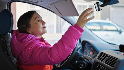 Mature hispanic woman with grey hair touching rearview sitting on car at street