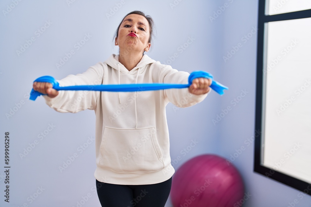 Poster middle age hispanic woman training arm resistance with elastic arm bands looking at the camera blowi