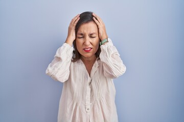 Middle age hispanic woman standing over blue background suffering from headache desperate and stressed because pain and migraine. hands on head.