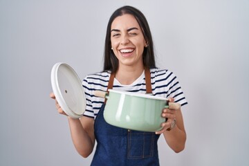Young brunette woman wearing apron holding cooking pot winking looking at the camera with sexy...
