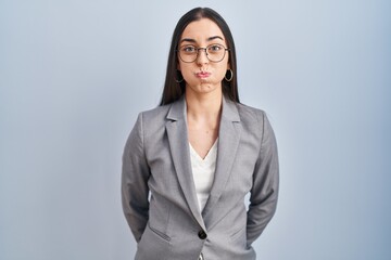 Hispanic business woman wearing glasses puffing cheeks with funny face. mouth inflated with air,...