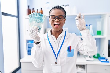 African woman with braids working at scientist laboratory holding money smiling and laughing hard out loud because funny crazy joke.
