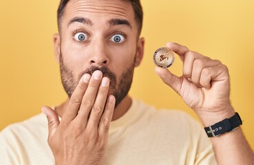 Handsome hispanic man holding uniswap cryptocurrency coin covering mouth with hand, shocked and...