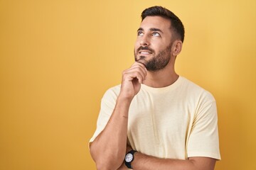 Handsome hispanic man standing over yellow background with hand on chin thinking about question, pensive expression. smiling and thoughtful face. doubt concept.