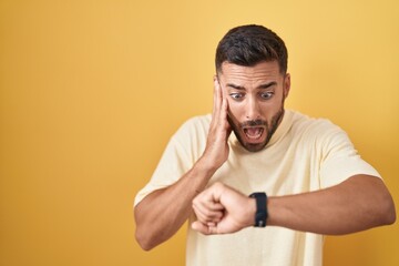 Handsome hispanic man standing over yellow background looking at the watch time worried, afraid of...