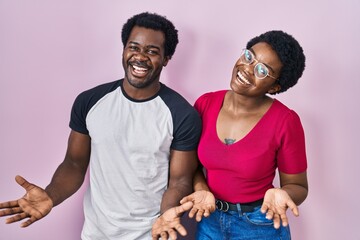 Obraz premium Young african american couple standing over pink background smiling cheerful with open arms as friendly welcome, positive and confident greetings