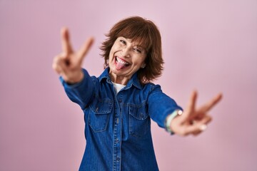 Middle age woman standing over pink background smiling with tongue out showing fingers of both...