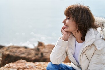 Middle age woman smiling confident sitting on the rock at seaside