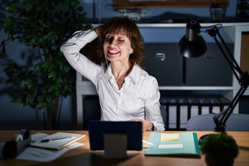 Middle age woman working at the office at night smiling confident touching hair with hand up gesture, posing attractive and fashionable