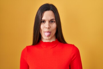 Fototapeta premium Young hispanic woman standing over yellow background sticking tongue out happy with funny expression. emotion concept.