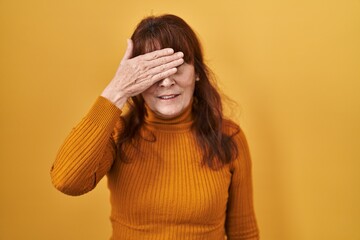 Middle age hispanic woman standing over yellow background covering eyes with hand, looking serious and sad. sightless, hiding and rejection concept