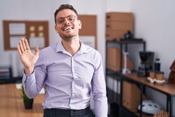 Young hispanic man at the office waiving saying hello happy and smiling, friendly welcome gesture