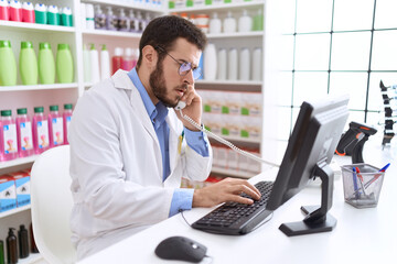 Young hispanic man pharmacist talking on telephone using computer at pharmacy