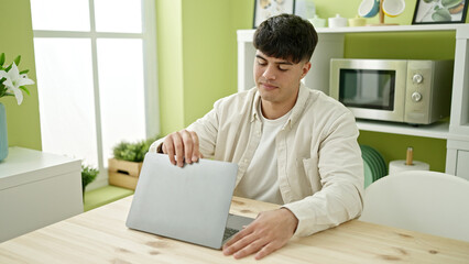 Young hispanic man opening laptop sitting on table at dinning room