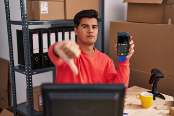 Hispanic man working at small business ecommerce holding dataphone with angry face, negative sign...
